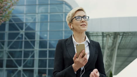 Sonrió-A-Una-Atractiva-Mujer-De-Negocios-Con-Gafas-Y-Auriculares-Paseando-Y-Relajándose-Mientras-Disfrutaba-De-La-Música-En-Su-Smartphone-Antes-De-Una-Reunión-Importante