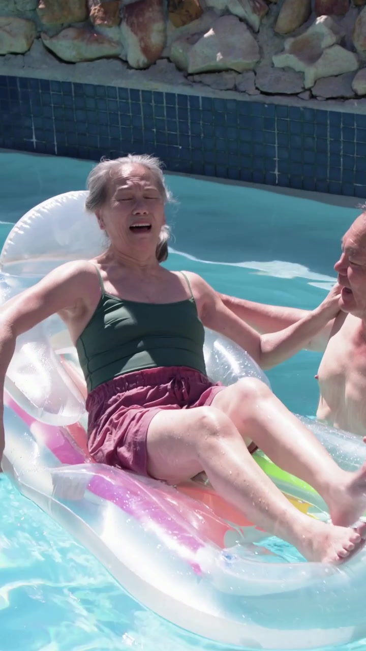 Vertical video: Shifting older woman reaching man entering pool by float in swimsuit seeking touch