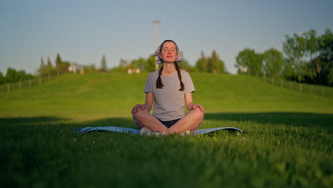 a smiling young woman sits on her yoga mat, glowing skin lit by the golden sun, with a green hill behind and a soft shadow dissolving across the grass. dolly right.
