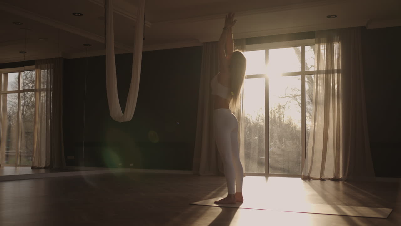 A young woman in white sportswear is stretching with a large hall with large windows in a slow-motion scheme the sun's rays shine through the window.