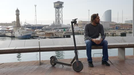 man takes a break from scooter trip and sits on bench sipping hot drink