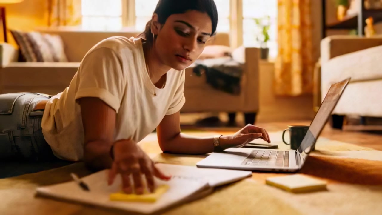 Starting study, woman typing on laptop reaching for sticky notes at home wearing tee and jeans