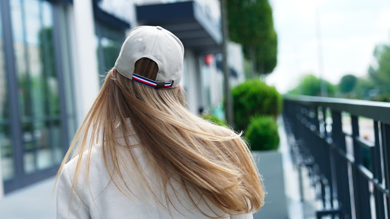 Blonde long-haired model turning in front of camera. Lady in jacket, cap and sunglasses demonstrating clothes in the street. Blurred backdrop.