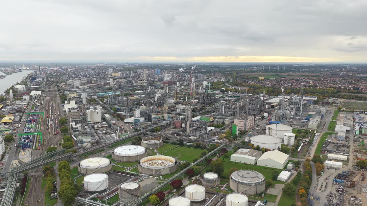 Vast industrial area featuring an oil refinery with large storage tanks, complex pipeline networks, and smoking chimneys near a major river