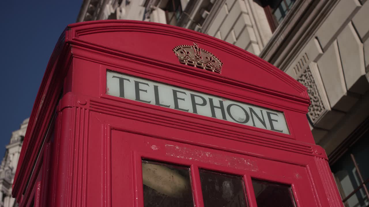 Bright red phone box with royal crown detail in front of historic city building, sunshine and blue sky highlighting classic London icon visited by many people each year