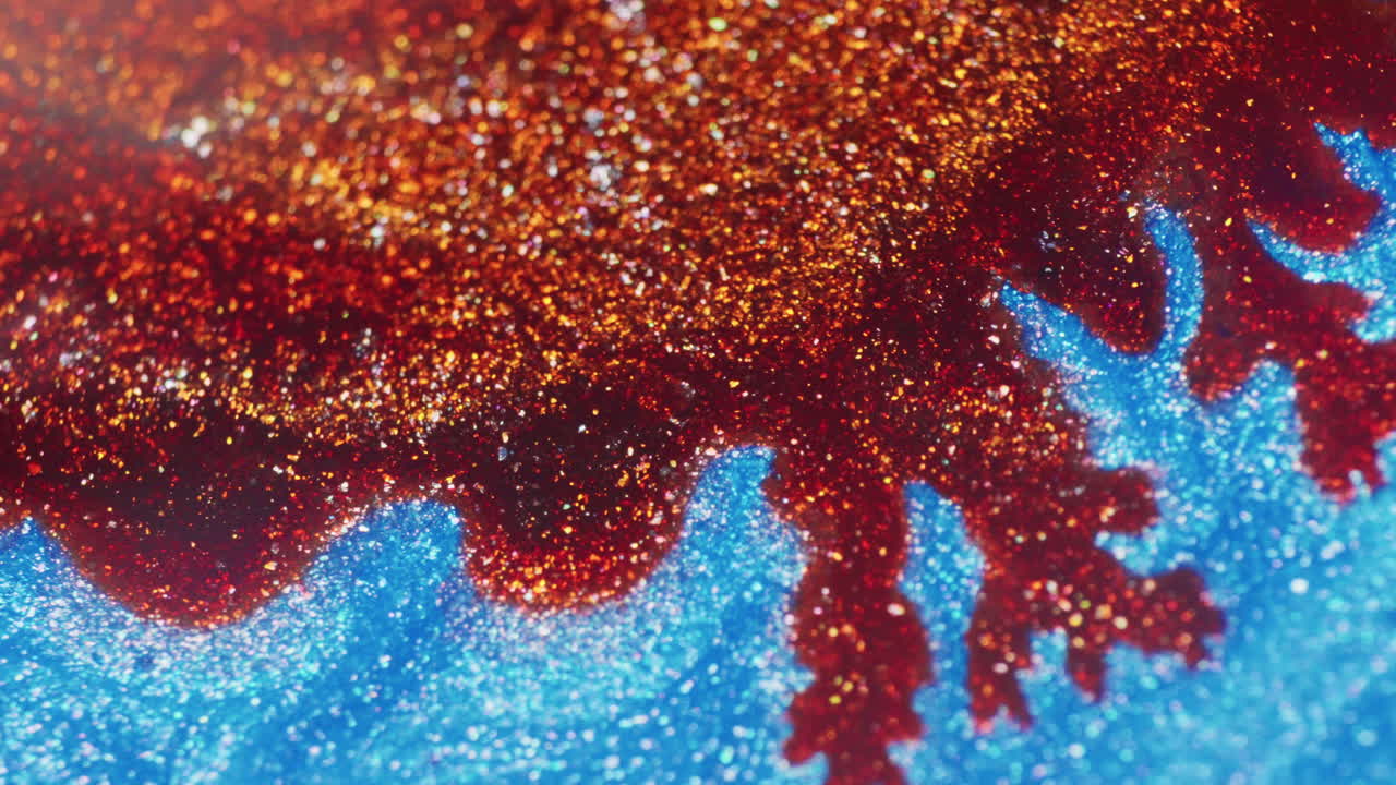 Macro shot of glittery red, bordeaux, and orange ink spreading across shimmering blue ink as liquid disperses on the surface