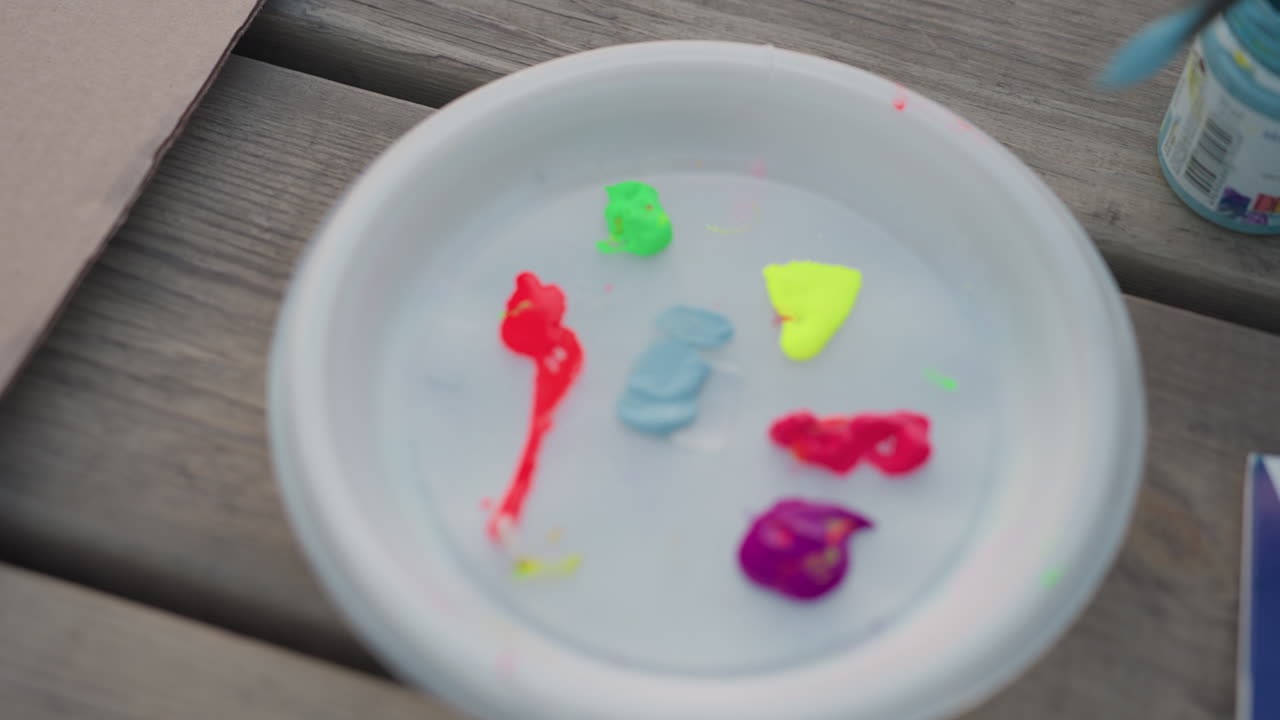 Prato plástico branco com manchas de tinta acrílica neon vermelha, verde, amarela, rosa e roxa sobre mesa de madeira envelhecida ao ar livre, enquanto pincel pega tom azul para misturar em atividade de arte criativa à beira do rio ao pôr do sol.