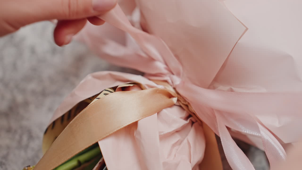 Mani che legano un nastro sul bouquet, fiorista che rifinisce l'incarto di carta pastello con un nastro di raso e un fiocco delicato, dettaglio in primo piano che mostra le dita che sistemano gli steli, illuminazione calda e soffusa, preparazione intima di un regalo.