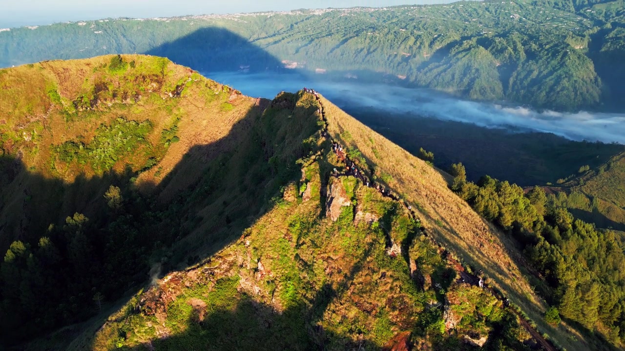 Sentiero su cresta vulcanica con escursionisti che camminano lungo una cresta esposta sopra una lontana foschia mattutina, luce dell'alba che modella il terreno accidentato e il paesaggio della caldera sul Monte Batur, ripresa aerea con drone in tilt-up.