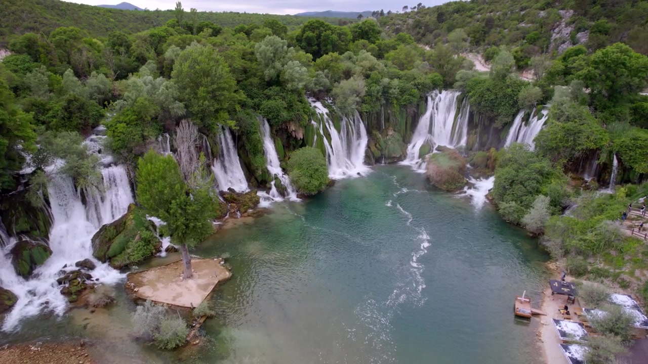 La cascata di Kravica circonda un'ampia pozza turchese con una fitta boscaglia verde in Bosnia, ripresa aerea con movimento a sinistra che stabilisce lo sfondo.