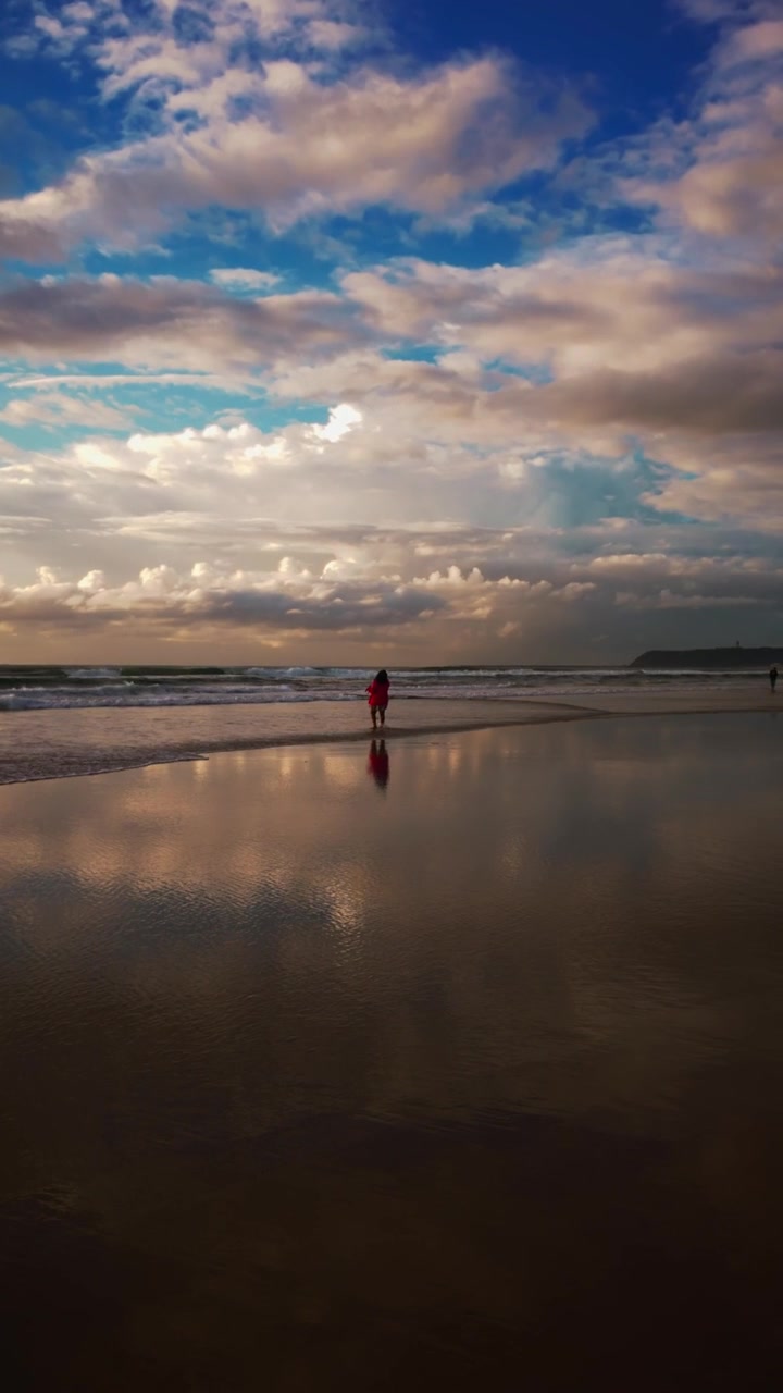 Vertikal video af en ensom skikkelse, der går langs den afsidesliggende Sandwood Bay strand i Skotland, med dramatiske skyer reflekteret i vådt sand. Levende himmel, blødt lys, vidvinkelkomposition.