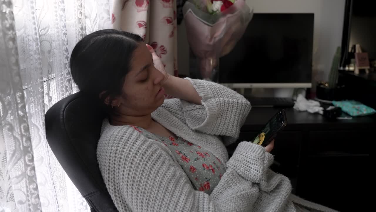 Giovane donna asiatica bengalese che controlla il suo telefono mentre è seduta su una sedia la mattina dopo essersi svegliata