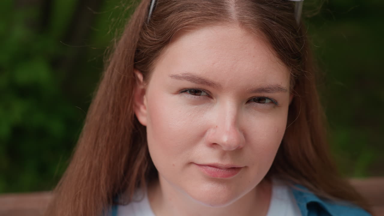 Close up of cute lady with long hair and white sunglasses on head sitting outdoors giving serious yet charming look, sunlight highlighting facial expression against blurred green background