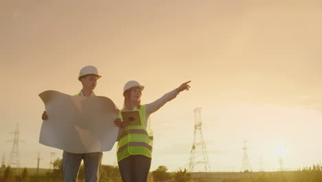 Zwei-Elektriker-Arbeiten-Zusammen-Und-Stehen-In-Helmen-Auf-Dem-Feld-In-Der-Nähe-Der-Stromübertragungsleitung.-Zwei-Elektriker-Arbeiten-Zusammen-Und-Stehen-Auf-Dem-Feld-In-Der-Nähe-Von-Strommasten.-Umweltfreundlicher-Kraftstoff