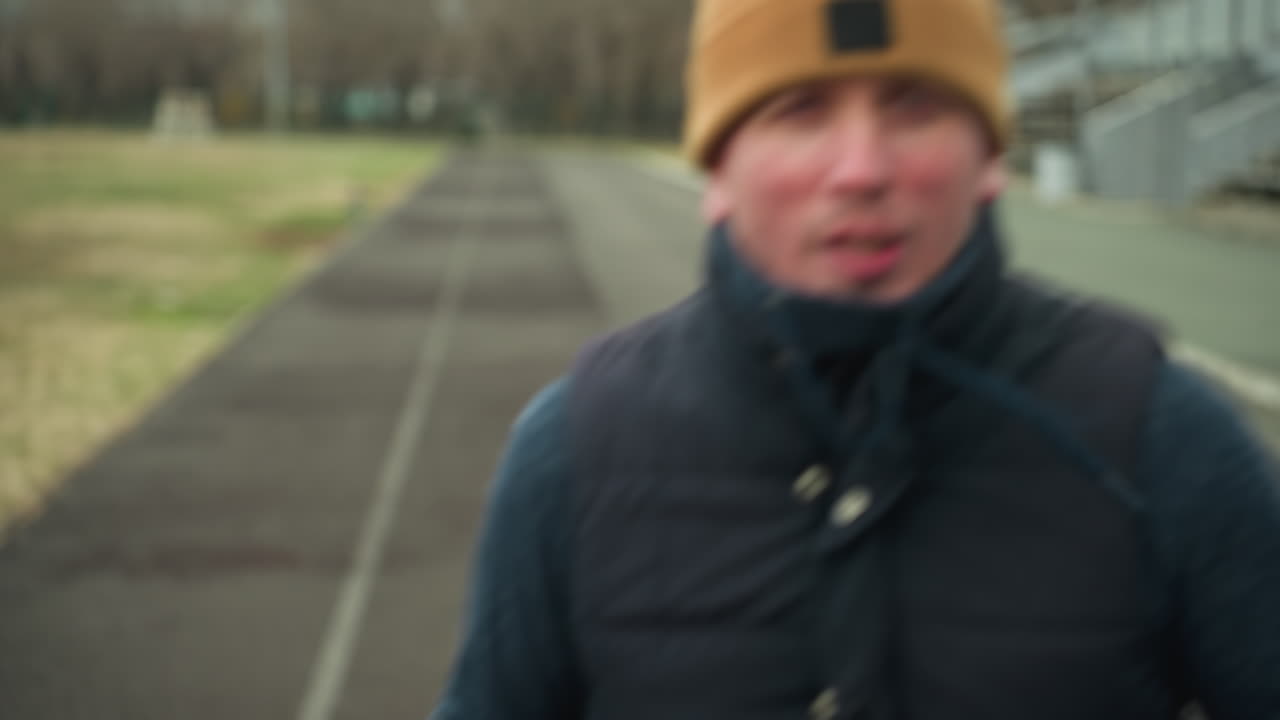 Close-up of a coach jogging on a track in a stadium, talking to himself, the blurred background shows a football field