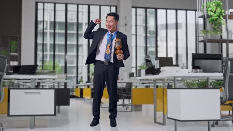 cuerpo lleno de hombre de negocios asiático en traje y corbata con una medalla de oro y trofeo flexionando su bíceps y sonriendo a la cámara en la oficina