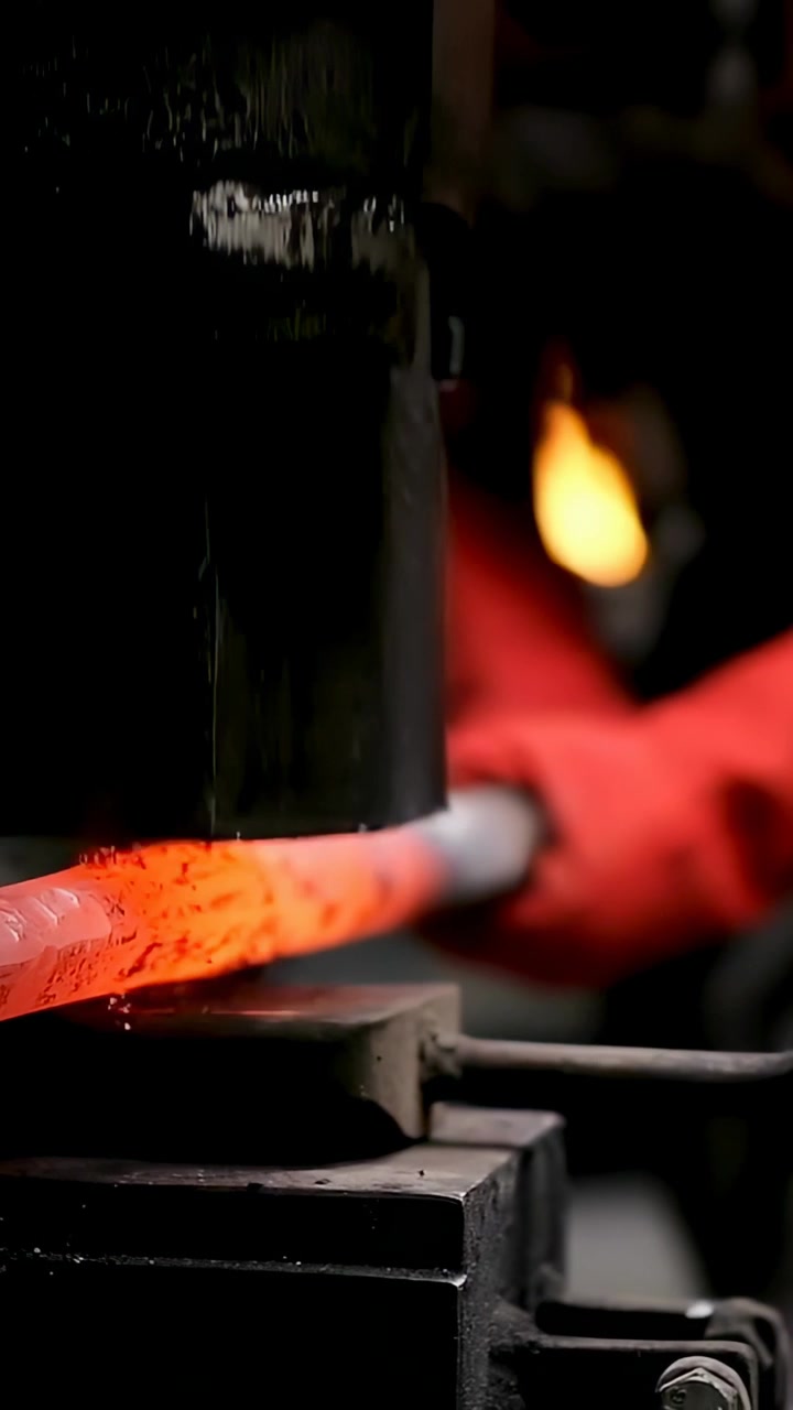 Vertical video: Smith in red gloves positioning hot rod under descending hammer in forge, forging