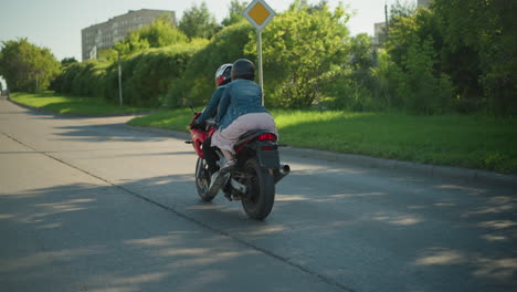 two ladies wearing helmets ride a red power bike through a shaded path lined with lush green trees, they approach a sunny path, with a blur view of car and building visible in the distance