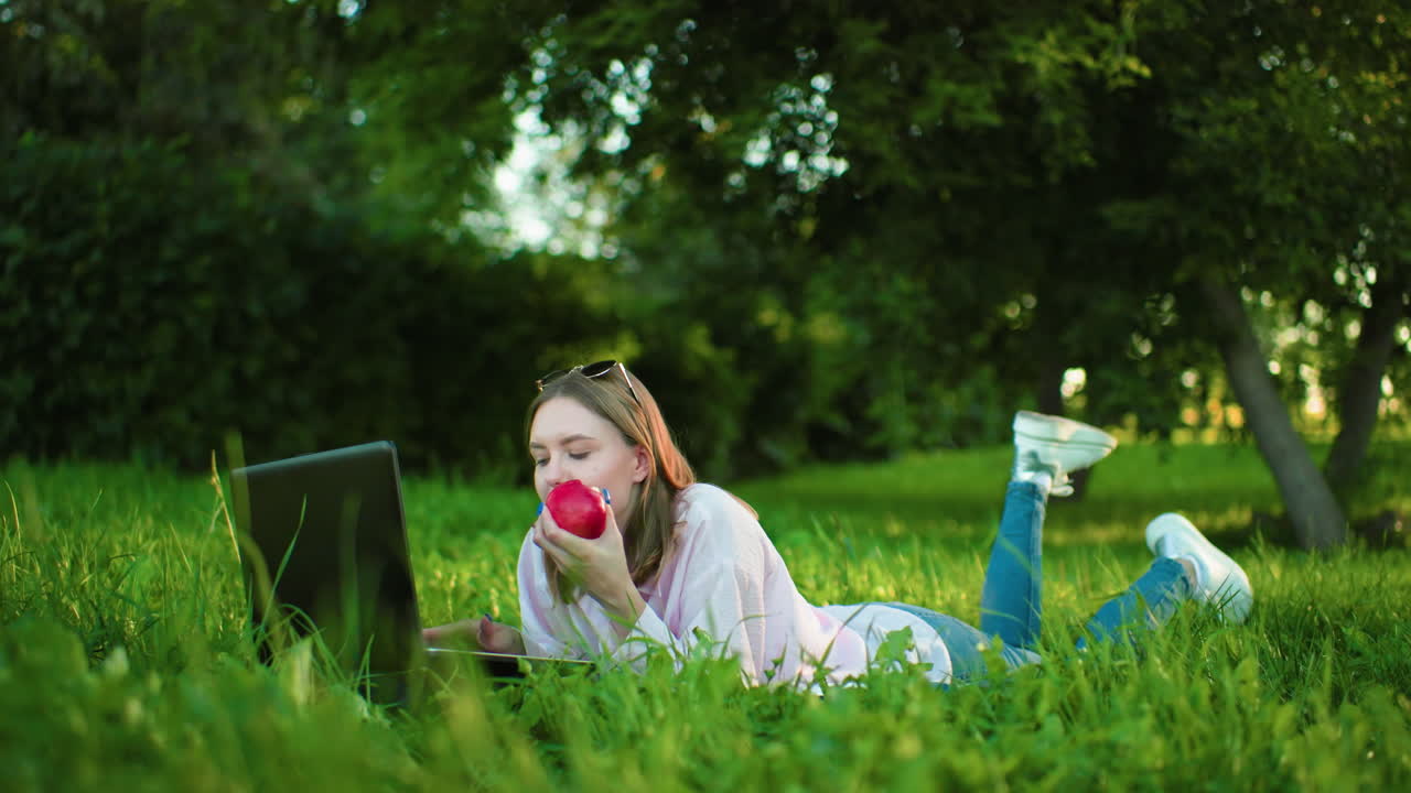 Freelancer lying on grass using laptop while eating apple and typing with leg slightly raised, surrounded by vibrant greenery and trees creating a relaxed outdoor work environment