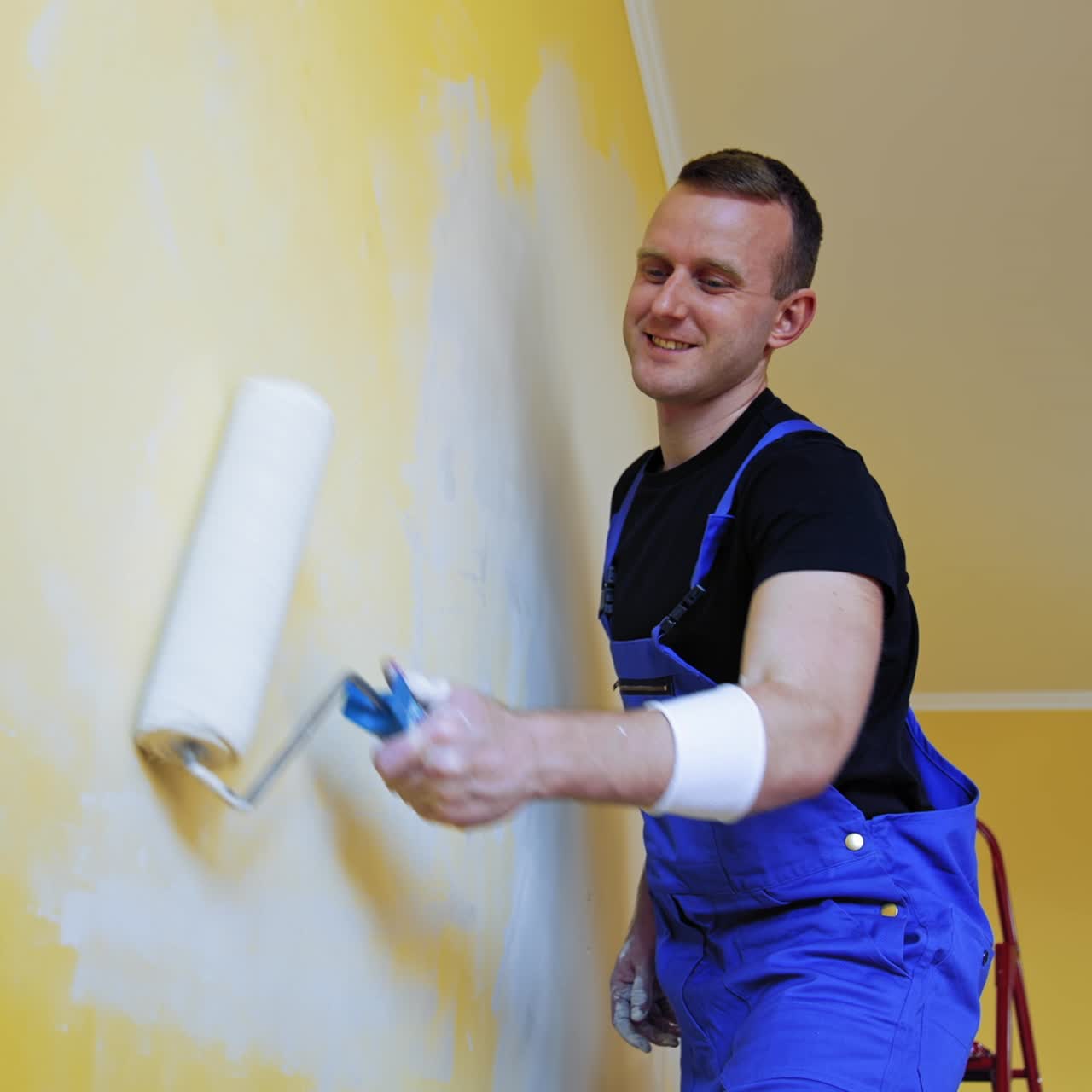 Young man improving apartment. Interior decoration in flat. Worker in blue overalls painting wall with a paint roller. Painting services