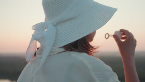 Side view of woman in white sun hat blowing soap bubble over river valley at sunset, tall grass framing scene, warm golden light highlighting profile and leisurely moment