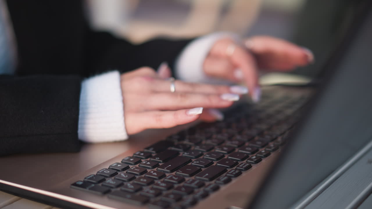 Close-up of hands with polished nails and delicate rings typing on laptop keyboard outdoors, placed on wooden table with blurred background