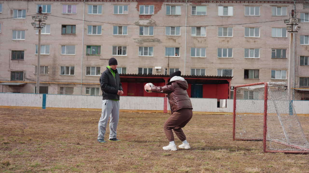 Вратарь отрабатывает технику с белым тренером и взрослой женщиной, сосредоточен на сейвах и стойке в приседе возле сетки, на заднем плане — фасад городского дома, пасмурное небо, утепленные куртки и кроссовки, повторение для развития рефлексов