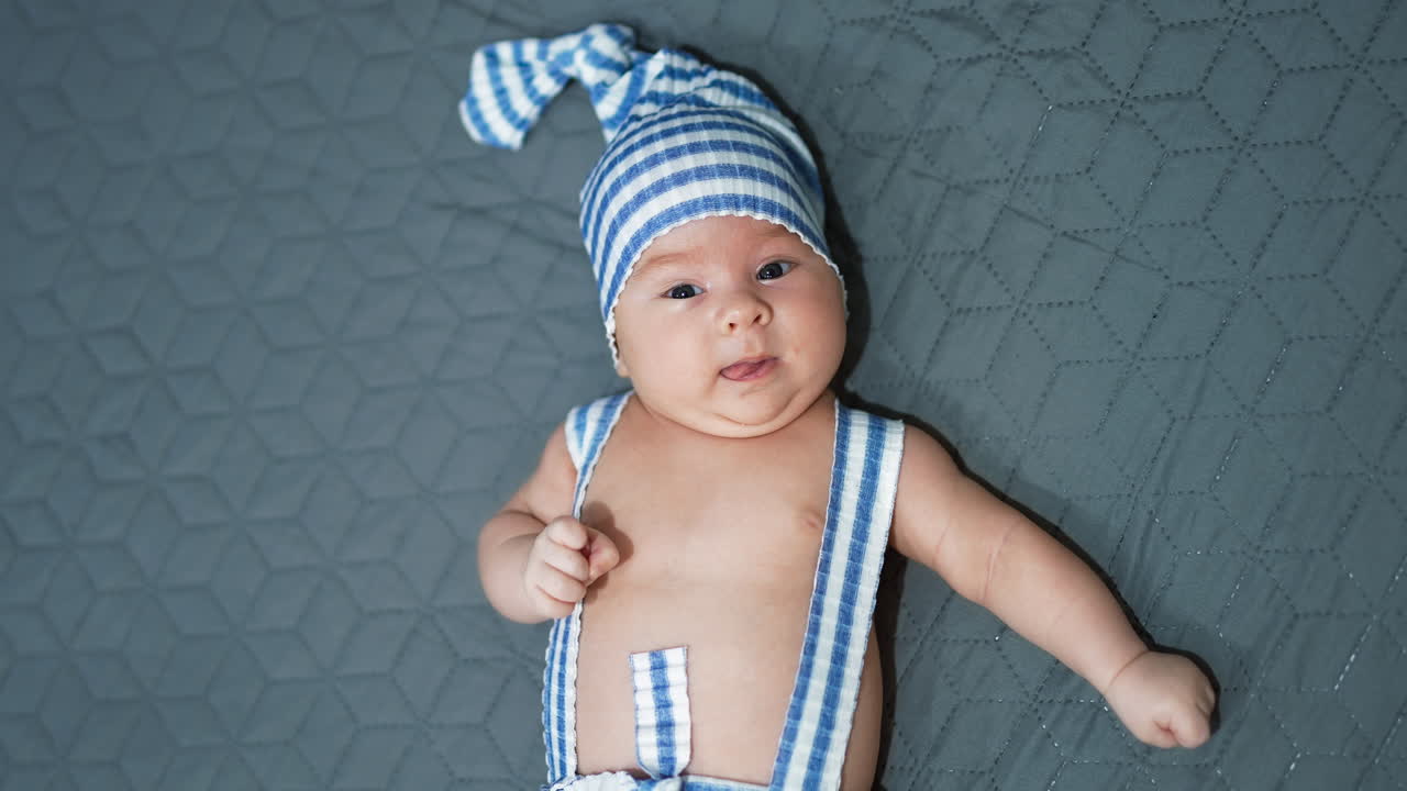 Sweet boy wearing striped suit lies on the bed with his hands sideways. Funny costume on a kid at the grey background.