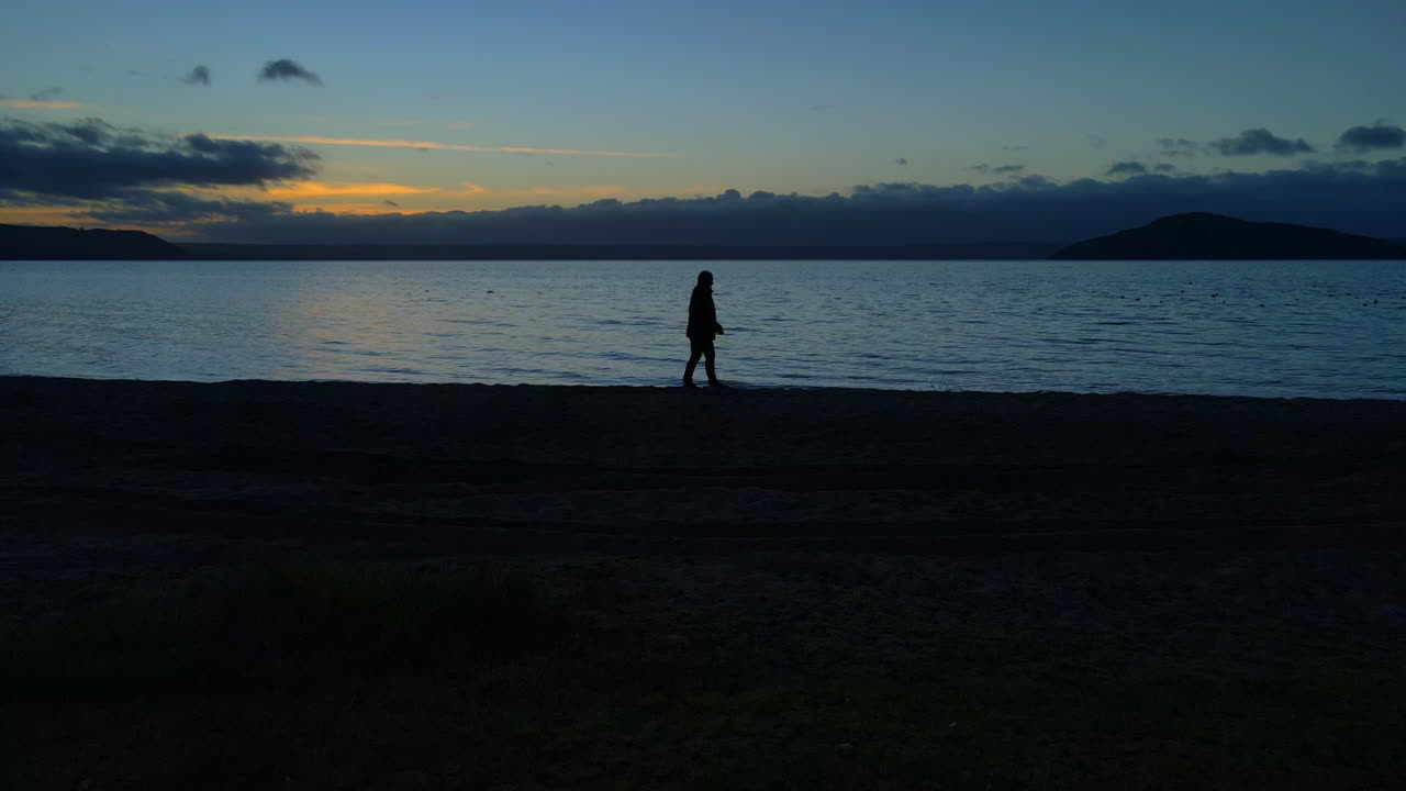 Uomo solitario che cammina lentamente da solo lungo la riva al tramonto con l'orizzonte sullo sfondo