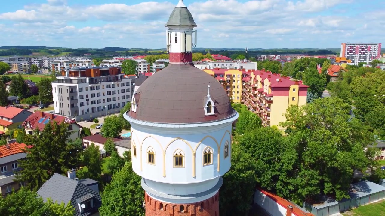 Luftopptak av vanntårnet i Ełk, Polen, med grønne trær og fargerike bygninger rundt, skyet, 33 meter høyt, med plass til tekst
