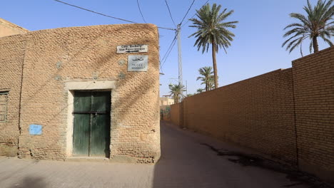 Sunlit-alley-in-Sbeitla-with-brick-buildings-and-palm-trees,-blue-sky