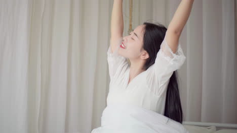 피자마를 입고 일어나는 아름다운 소녀의 실제, 큰 창문 근처에 앉아있는 침대에서 깨어나는, 완전히 휴식을 취하고, 아침에 신선한 공기와 빛을 얻기 위해 커튼을 펴는.