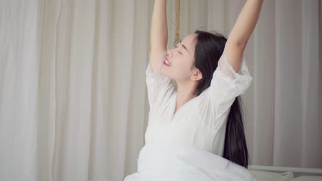 real shot of beautiful girl getting up in white pajamas waking up in her bed sitting near the big window fully rested and open the curtains stretching in the morning to get fresh air and sunshine