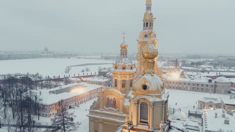 aerial footage of the peter and paul fortress in a snow storm in a winter evening, night illumination, petropavlos cathedral shines with golden light, sights of st. petersburg in the background