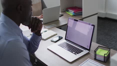 African-american-businessman-sitting-at-desk-and-using-laptop-with-copy-space-on-screen-in-office