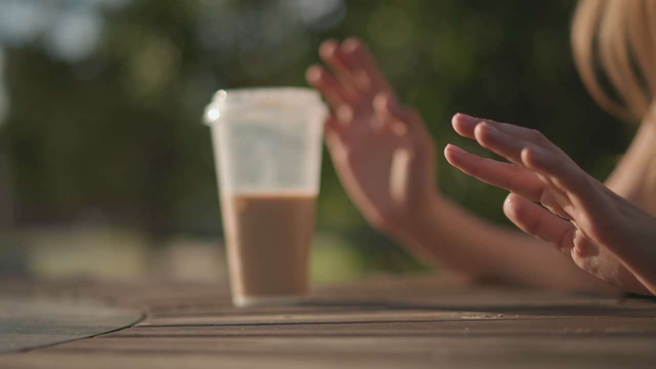 primo piano di mani accanto a una tazza di caffè freddo su un tavolo di legno illuminato dal sole, dita che tamburellano e gesticolano in slow motion, latte cremoso in un bicchiere di plastica trasparente, profondità di campo ridotta con alberi bokeh sfocati