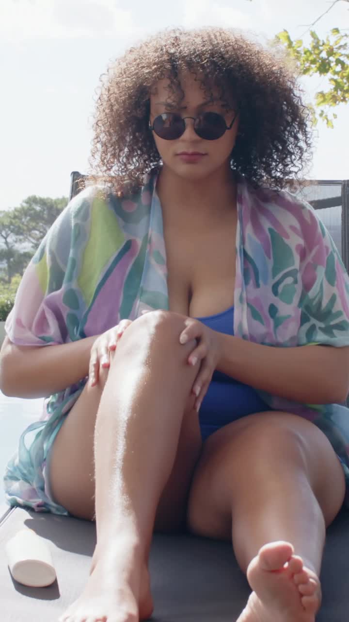 Vertical video: Sitting woman in shades rubbing lotion on legs at patio for sun care, with swimsuit