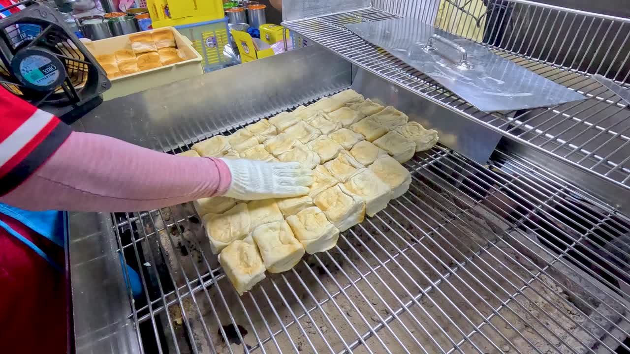 Gloved vendor grills bread cubes on metal grate at bustling, brightly lit Bangkok street market