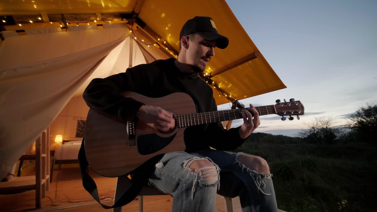Mand spiller guitar og synger sange i et telt med lyspærer.