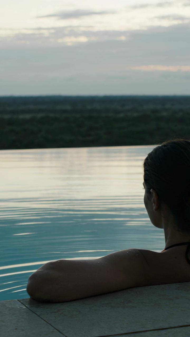 Vertikales Video: In der Dämmerung bewegt sich eine Frau im Badeanzug am Rand eines Infinity-Pools, blickt zum Horizont, mit Platz für Text