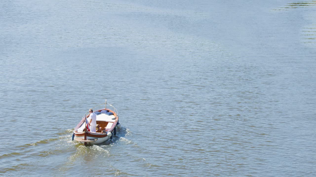 Aerial view of two tourists rowing a small boat across a calm river under bright daylight, with gentle ripples trailing behind. Steady camera, tranquil mood