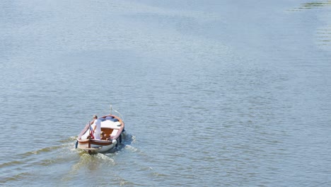 aerial view of two tourists rowing a small boat across a calm river under bright daylight, with gentle ripples trailing behind. steady camera, tranquil mood