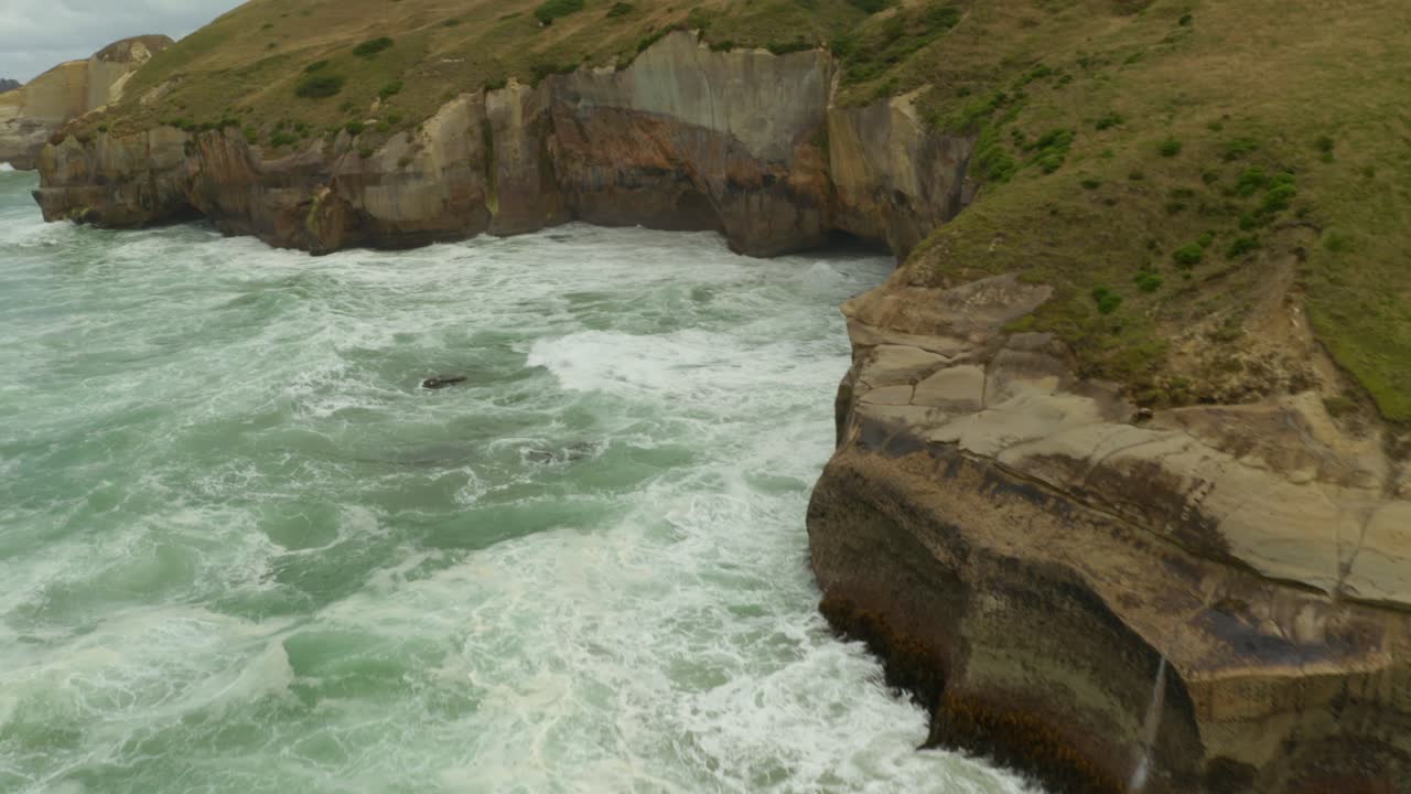 낮 동안 뉴질랜드의 절벽 벽에 부딪히는 파도와 함께 해안선을 따라 절벽의 공중 패닝 및 틸팅 샷