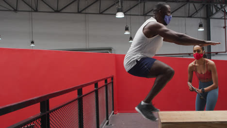 African-american-man-jumping-on-box-wearing-face-mask-at-gym,-caucasian-woman-motivating