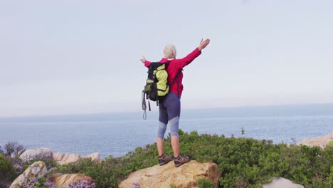 rear view of senior hiker woman standing with her arms wide open standing on a rock while trekking i