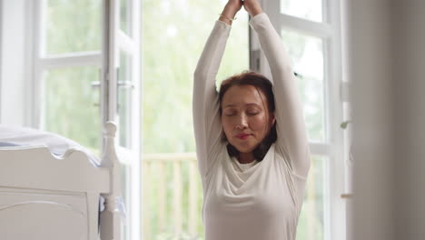 Mujer-Asiática-Madura-En-Pijama-Sentada-En-El-Suelo-Del-Dormitorio-Meditando-En-Pose-De-Yoga---Filmada-En-Cámara-Lenta