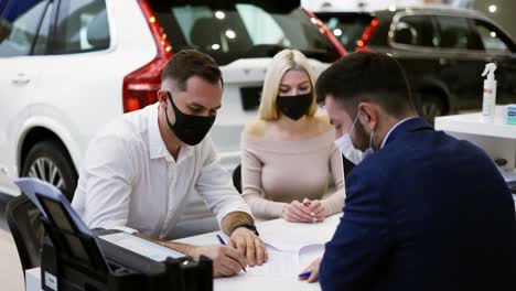 Pareja-Con-Máscaras-Compra-De-Un-Coche-Nuevo,-Firmando-El-Contrato-En-La-Sala-De-Exposición