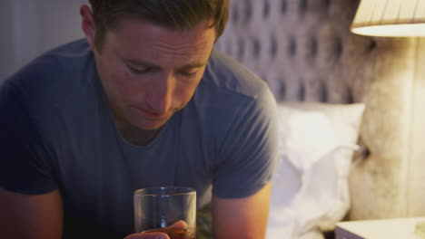 depressed man wearing pajamas sitting on side of bed with glass of whisky on bedside cabinet