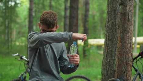 young boy wearing glasses and grey long-sleeve top opens bottle of water take a sip, while taking a break in a forest, bicycle is parked in the background, surrounded by tall green trees