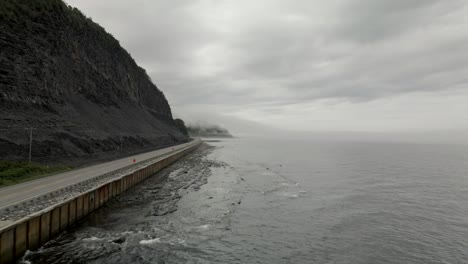 Flying-Over-Calm-Ocean-on-a-Cold-Morning-by-an-Empty-Road-Sitting-Below-a-Rocky-Mountain,-Drone,-Gaspesian